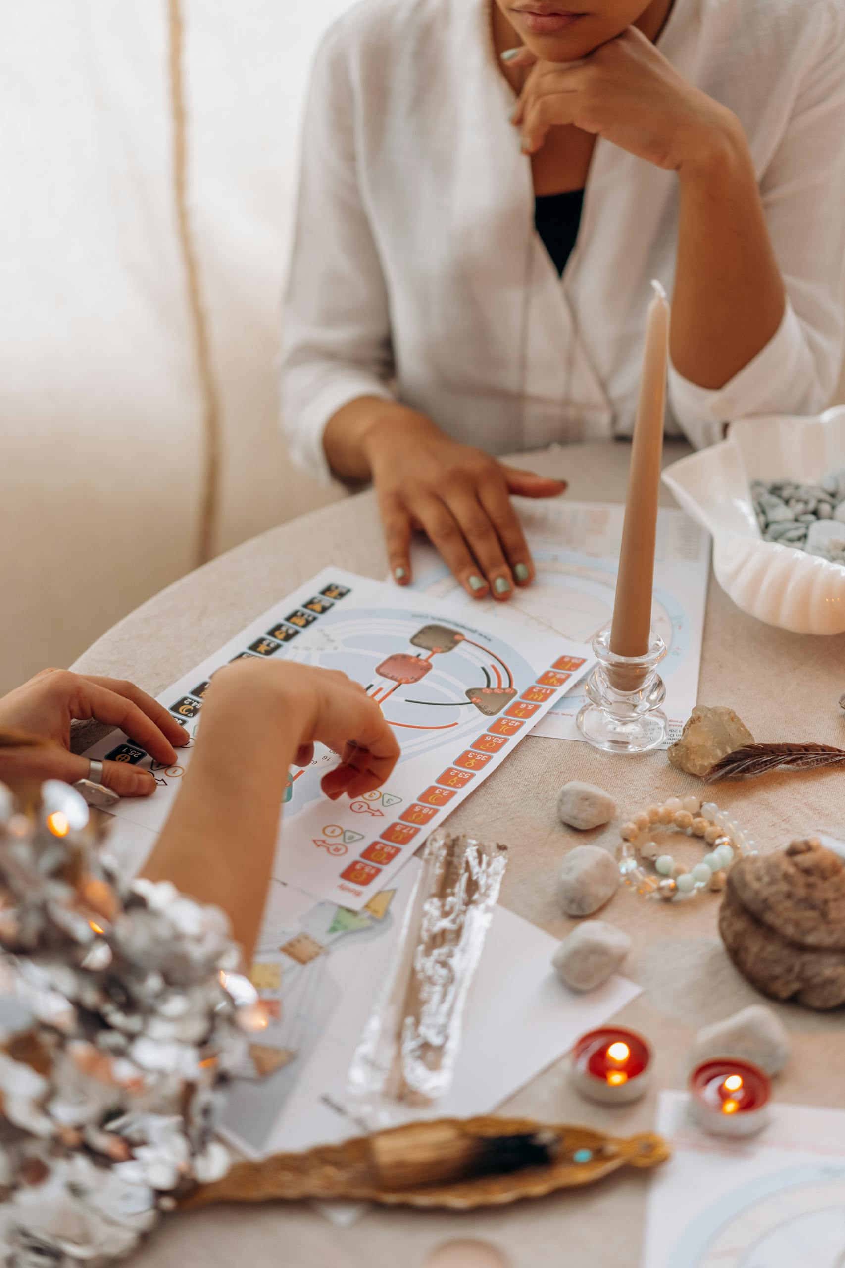 Two women engaged in a spiritual practice with candles, crystals, and illustrations on a table.